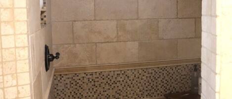 A narrow shower area with tiled walls, a tiled floor, a showerhead, and a small wooden bench.
