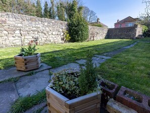Un mur de pierre, une cour gazonnée et un jardinier en bois rempli de plantes.