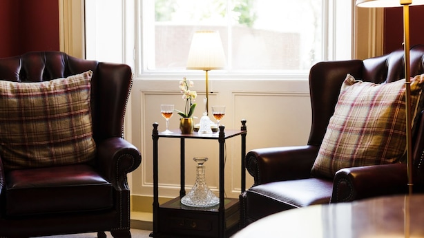 A room with two leather armchairs, a small table with a lamp, and a window with a view of greenery.