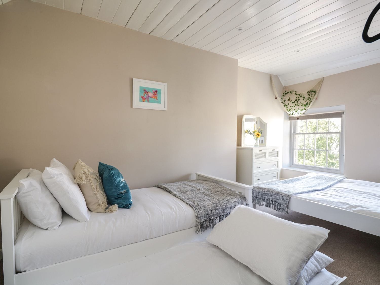 Quarto com cama branca, penteadeira branca, um banquinho e janela com vista para árvores.