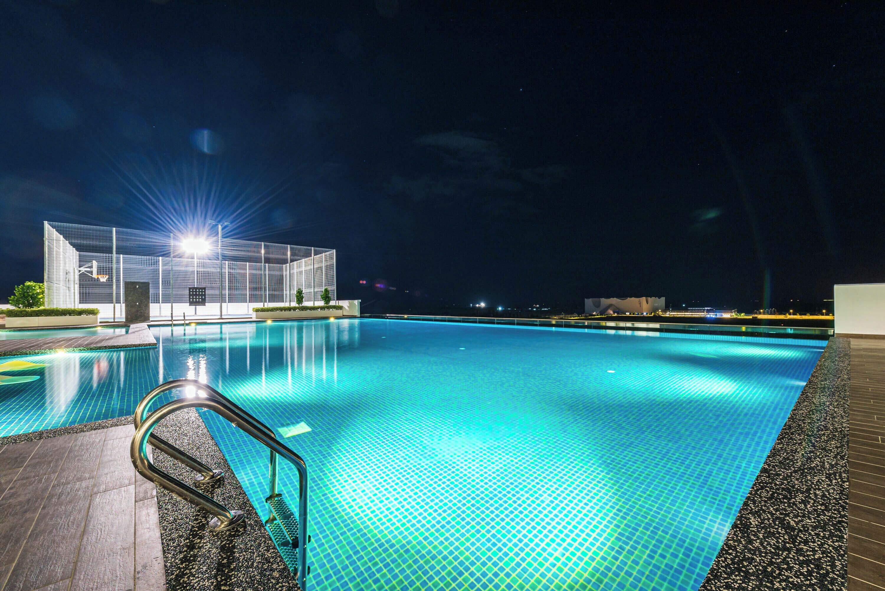 Ein Schwimmbecken mit Metallleiter, umgeben von einer gefliesten Terrasse, und ein Gebäude mit Glasfassade im Hintergrund.