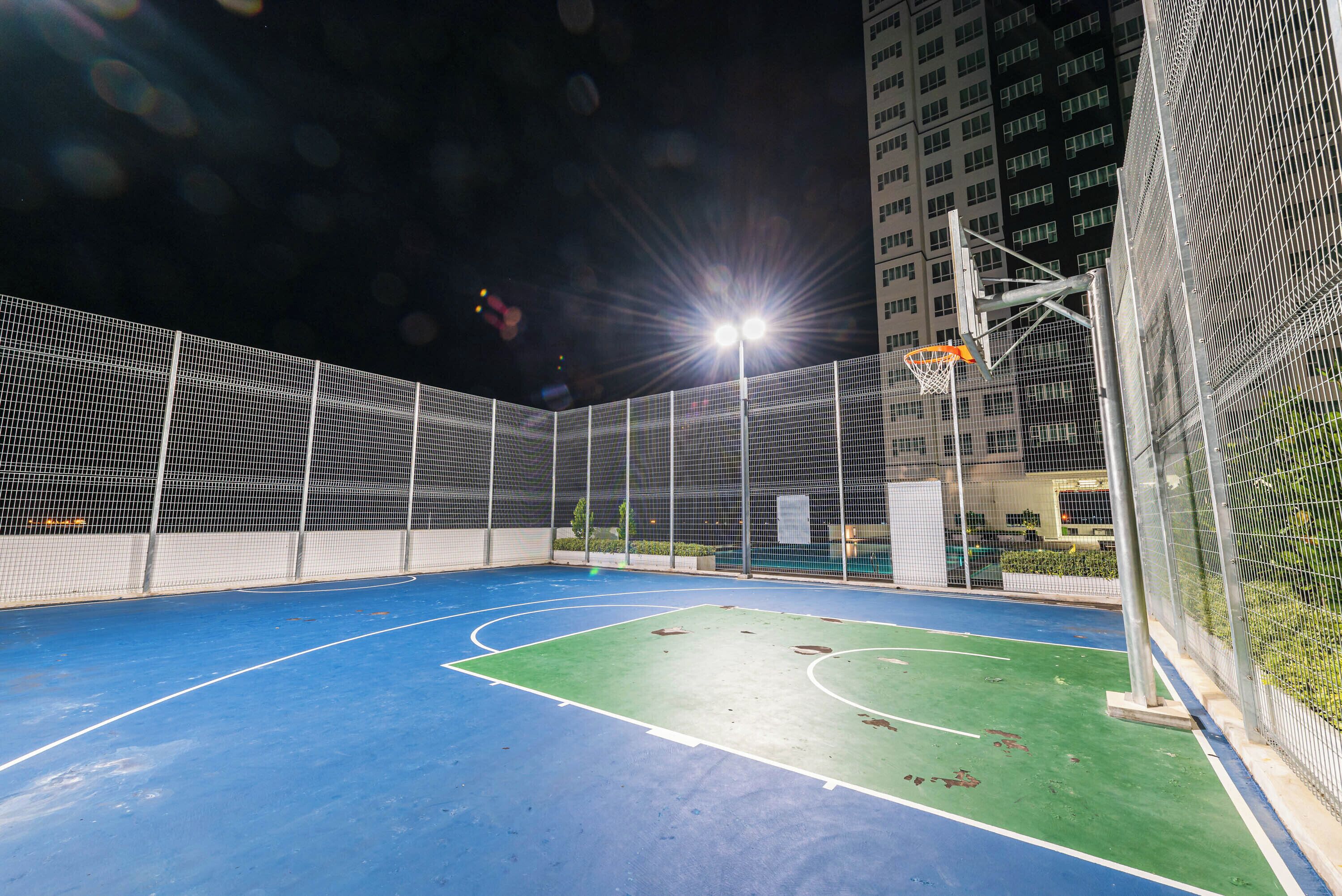An outdoor basketball court with a blue playing surface and a green area, surrounded by a metal fence.