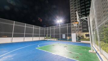 An outdoor basketball court with a blue playing surface and a green area, surrounded by a metal fence.