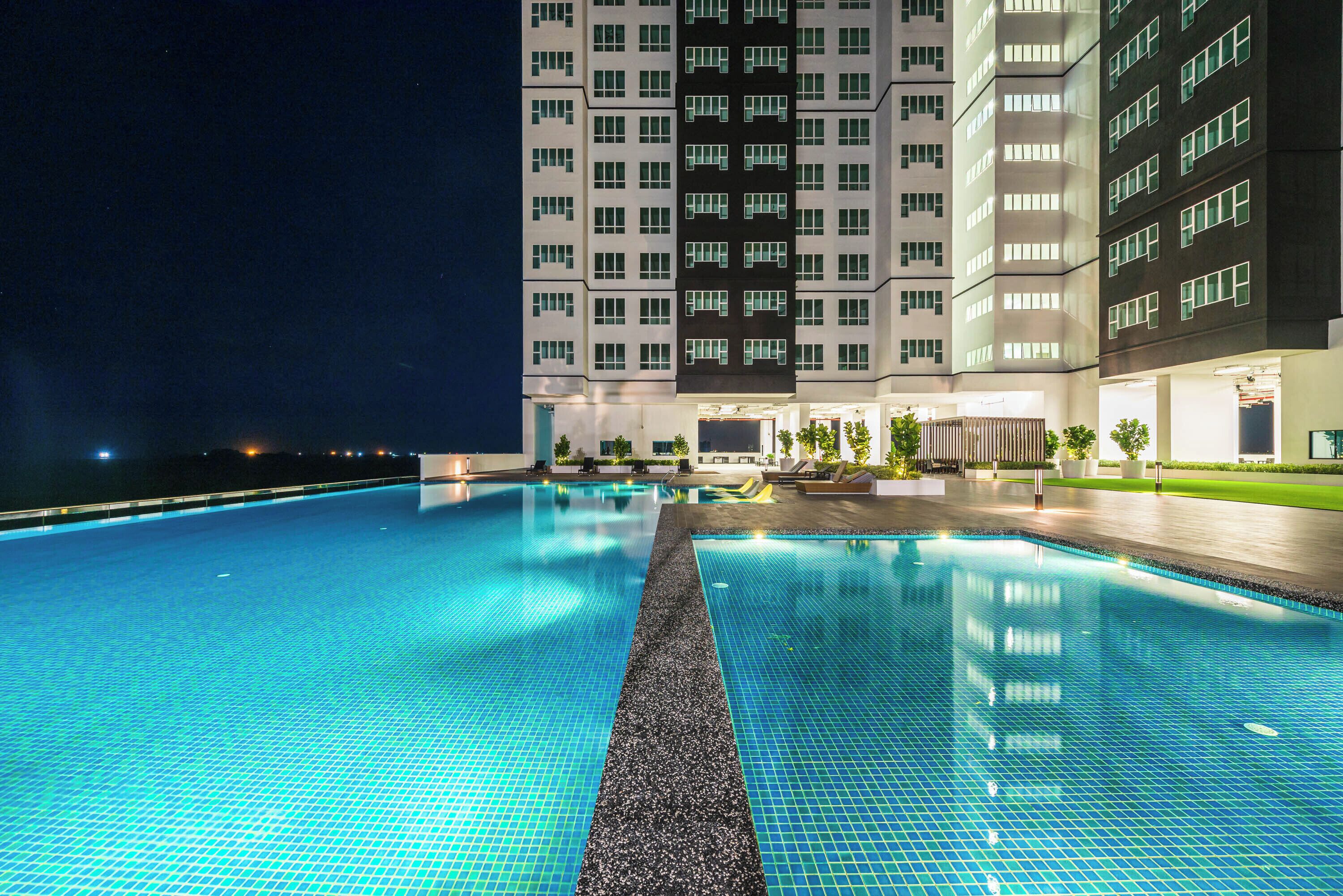 Una piscina con borde de azulejos, rodeada por un edificio de varios pisos y ventanas.
