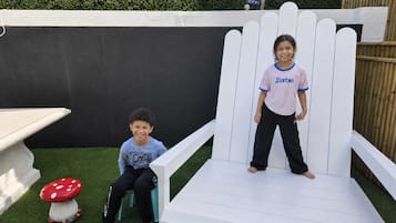 Two children sitting on a white bench in a garden with artificial grass and a decorative mushroom.