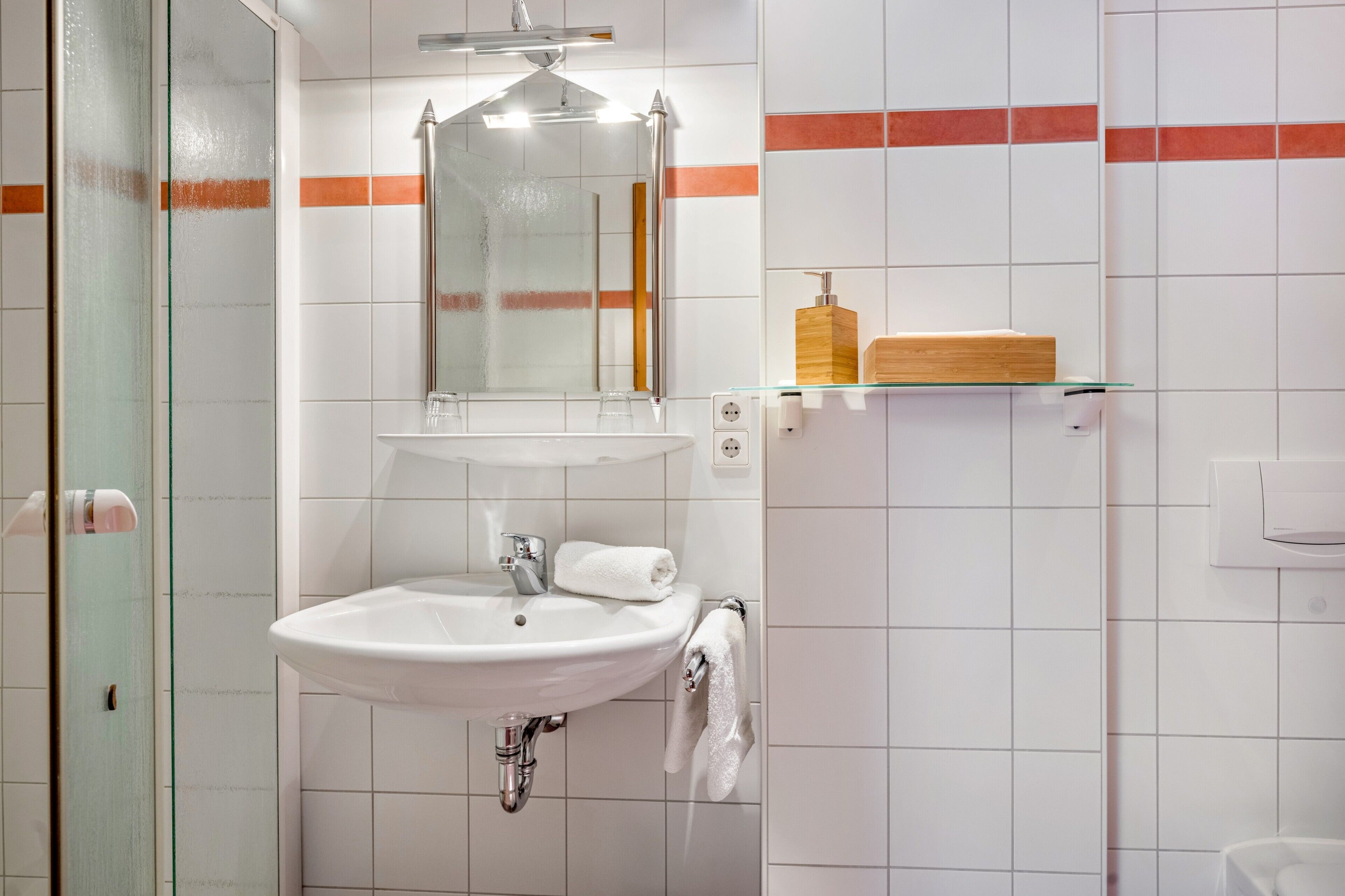 A bathroom with a glass shower enclosure, a sink with a mirror, and a wooden soap dispenser on a shelf.