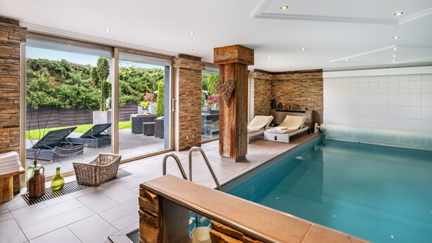 Una piscina interior moderna con pared de piedra, un área de descanso con un sofá y una silla, y vistas a un jardín a través de amplios ventanales.