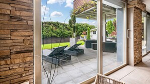 Un patio moderno con mobiliario exterior, un muro de piedra y un ventanal de vidrio que da a un jardín.