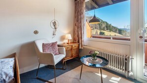 Una habitación con techo de madera, pared blanca, ventana con vistas a la montaña, armario de madera, mesa redonda con una lámpara y una silla con cojín.