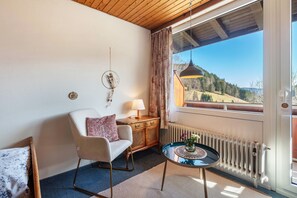 A room with a wooden ceiling, a white wall, a window with a view of a mountain, a wooden cabinet, a round table with a lamp, and a chair with a cushion.