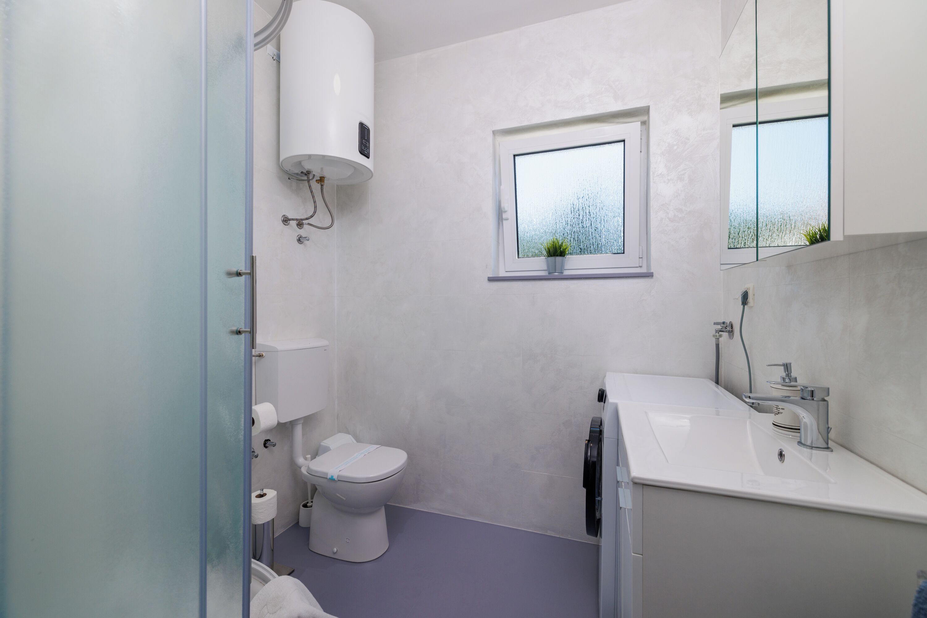 A bathroom with a toilet, a sink, a window with a plant, and a water heater.