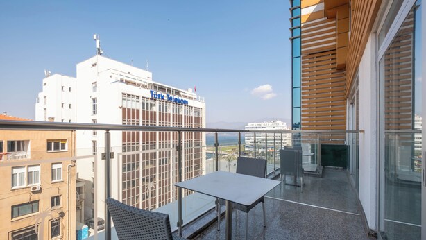 Balcón con una mesa y sillas, barandilla de vidrio y vista a un edificio con señalización de 'Turk Telekom'.