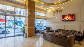 Un hall de hotel con un sofá grande, una araña, un televisor mostrando una chimenea y ventanas amplias.