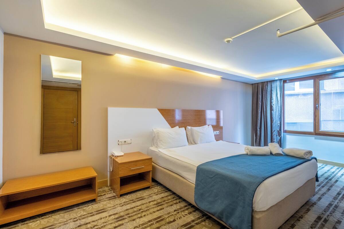 A hotel room with a bed, bedside table, mirror, and a window with curtains.