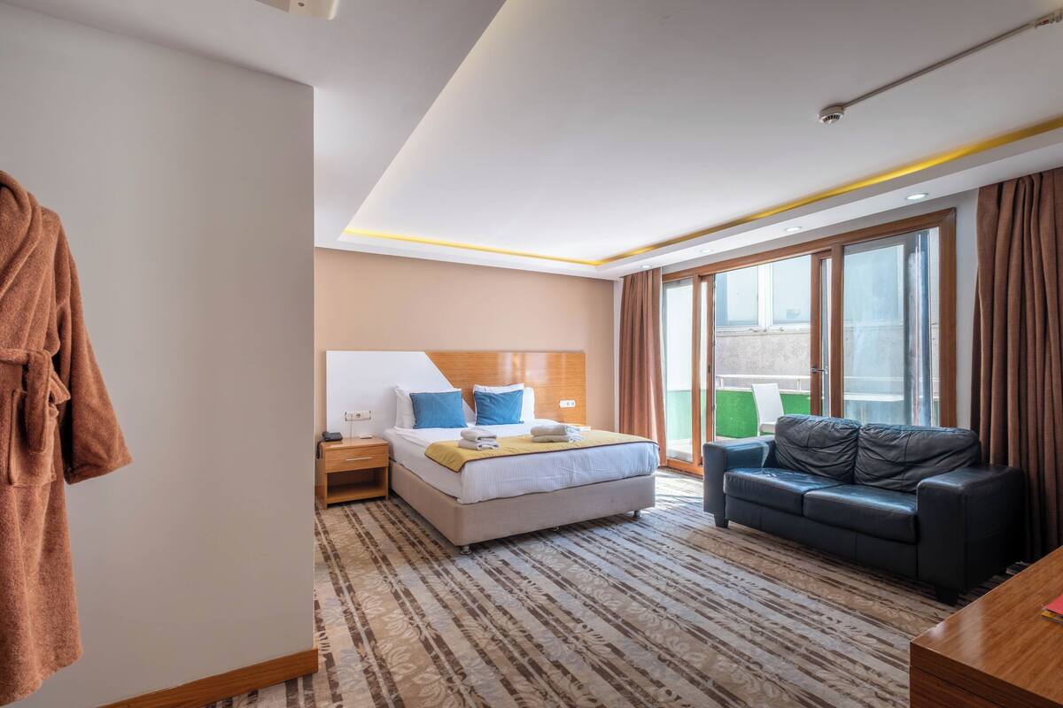 A hotel room with a bed, a sofa, a robe, a nightstand, and a large window.