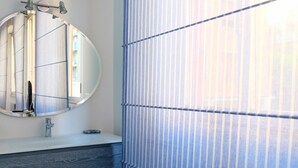 A bathroom with a round mirror, a sink, and a window with vertical blinds. - Hosmate - Milano - Vespri Siciliani- Blu Mare (Milano)