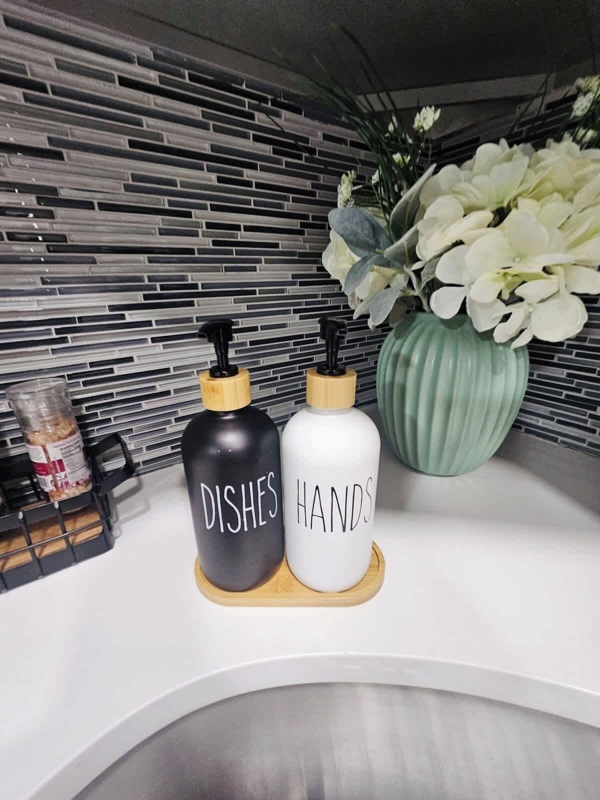 Un plan de travail de cuisine avec deux distributeurs de savon étiquetés « VAISSELLE » et « MAINS », un vase vert avec des fleurs blanches et un r écipient noir contenant un pot.
