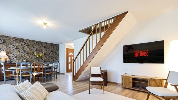 Una sala de estar moderna con una escalera, una zona de comedor y un televisor de pantalla plana mostrando 'Welcome to REIMS'.