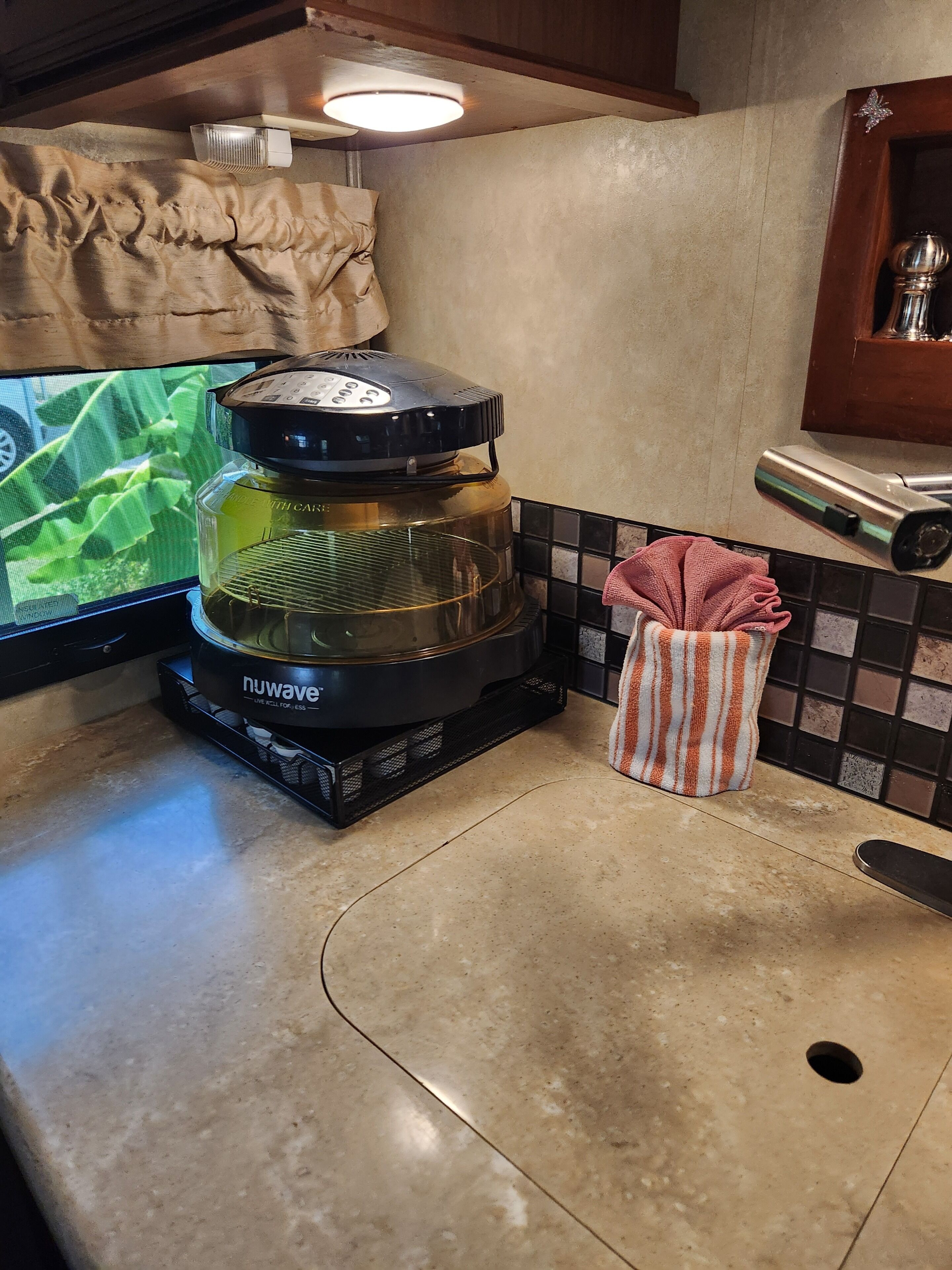 A kitchen with a Nuwave coffee maker, a television displaying a green screen, a tiled backsplash, and a small trash bin.