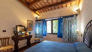 A bedroom with a bed, a wooden chair, a dresser, a mirror, and a window with floral curtains. - Tenuta Donna Vittoria Bouganville Apartment with Mountain View, Wi-Fi & Air Conditioning (Trapani)