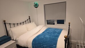A bed with white bedding and a blue blanket, a bedside table with a blue item, a wall clock, and a window with a blind. - Stylish Stay in Brighton: London Road Gem (Brighton and Hove)
