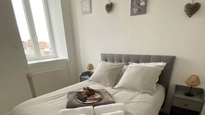 Una cama bien tendida con sábanas y almohadas blancas, una mesita de noche con una lámpara, y una ventana con vistas a edificios.