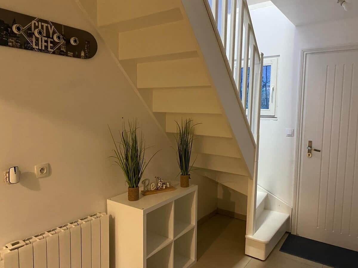 Un escalier avec une rampe blanche, une console blanche ornée de plantes, un skateboard sur lequel on peut lire le texte « CITY LIFE », et une porte avec une fenêtre.