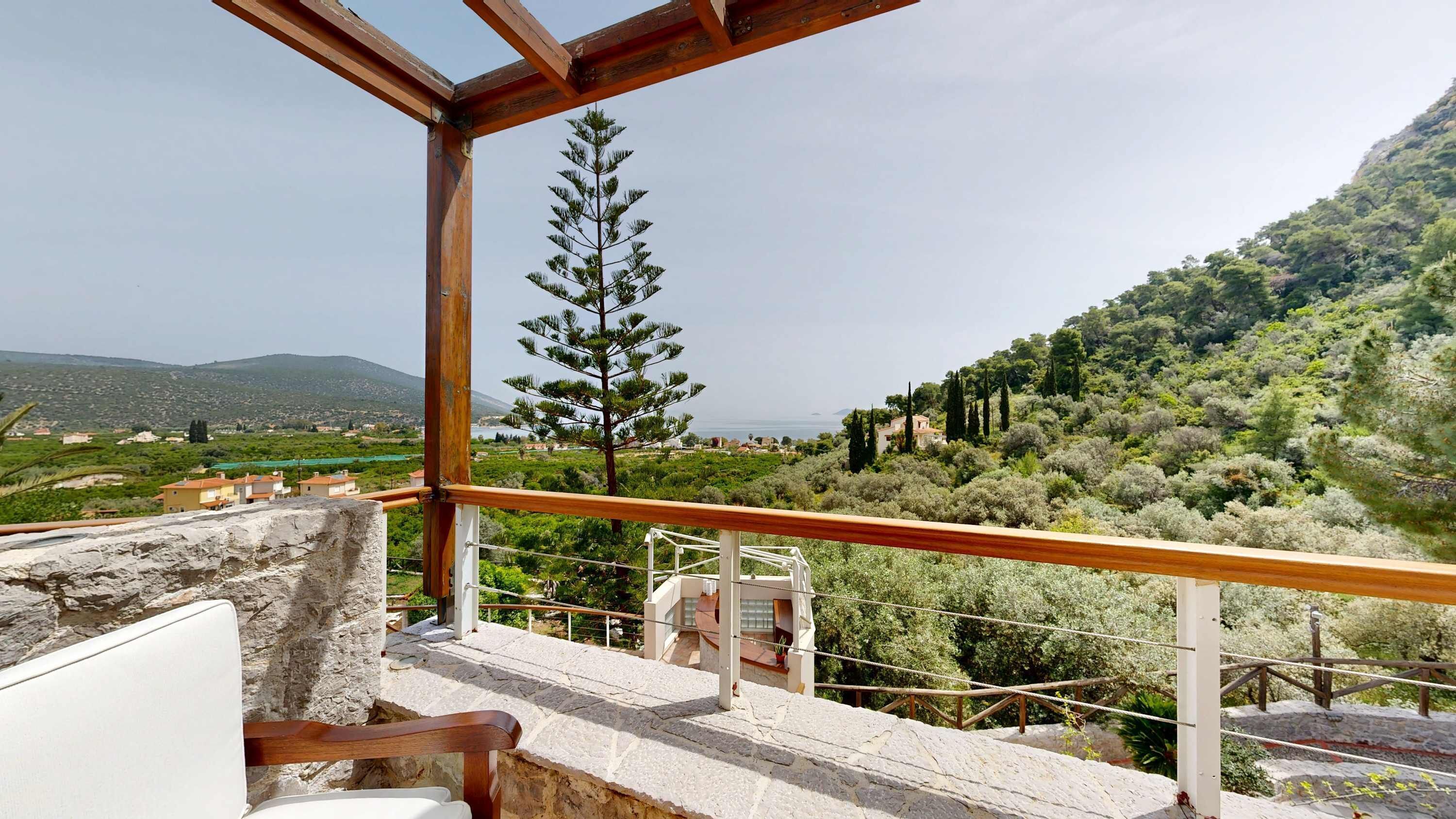 A balcony with a stone wall, a wooden railing, and a view of a mountainous landscape with trees and buildings.