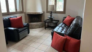 Ein Wohnzimmer mit Kamin, zwei Ledersofas mit roten Kissen, einem kleinen Tisch mit Lampe und großen Fenstern.