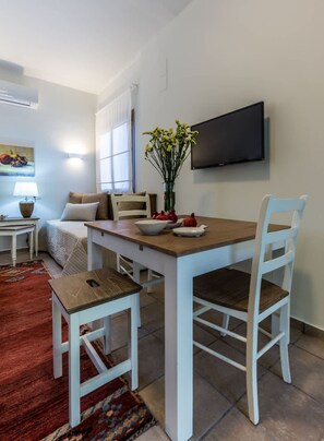 Comedor con mesa de madera y sillas blancas, un jarrón con flores, televisor en la pared y una cama con ventana al fondo.