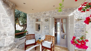 A stone-walled room with two wooden chairs and a small table, a door with glass panels, and a balcony with a stone balustrade. - Epidavros Villas - Elia Garden Retreat w Pool (Epidaurus)