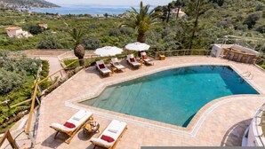 A pool area with sun loungers and umbrellas, surrounded by greenery and a view of the sea. - Epidavros Villas - Voukamvilia Retreat w Pool (Epidaurus)