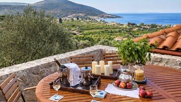 A round wooden table with chairs, candles, and a vase with flowers, set against a scenic view of the sea and hills.