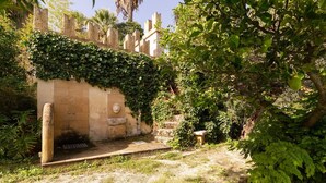 Un muro de piedra con una estatua, un escalón que lleva a una puerta y abundante vegetación.