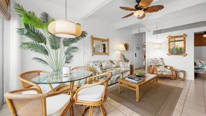 A spacious living area with a glass dining table, wicker chairs, a sofa with a tropical print, a ceiling fan, and a large potted plant. - Kona Mansions#B217 2nd Floor, Beautiful unit/Ocean views & Air Conditioning (Kailua Kona)