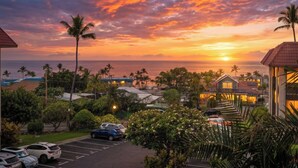 A coastal hotel with a view of the sunset over the ocean, a parking lot, and palm trees. - Kona Mansions#B217 2nd Floor, Beautiful unit/Ocean views & Air Conditioning (Kailua Kona)