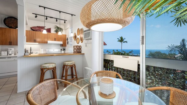 A modern kitchen with wooden cabinets, a glass dining table, wicker chairs, and a view of the ocean. - Kona Mansions#B217 2nd Floor, Beautiful unit/Ocean views & Air Conditioning (Kailua Kona)