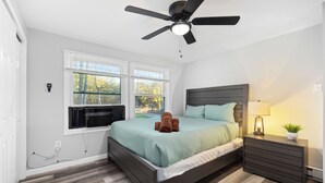 Un dormitorio con cama, ventilador de techo, mesita de noche con lámpara y ventana con persianas.