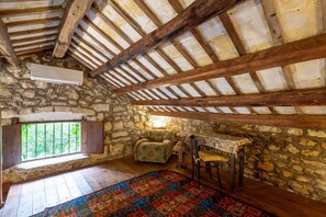 A rustic room with a stone wall, wooden beams, a window with bars, a patterned rug, a chair, and a table.