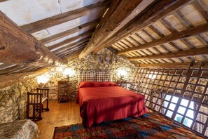 A rustic room with a stone wall, wooden beams, a bed with a red blanket, a wooden chair, and a patterned rug.