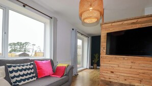 Un salon moderne avec un canapé gris, des coussins colorés, un grand téléviseur et un mur d’accent en bois.