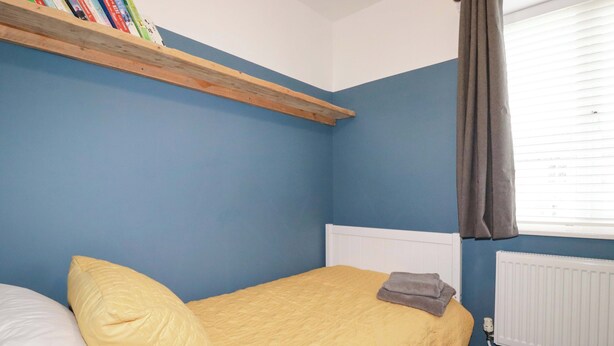 Une chambre à coucher avec un lit, des oreillers, un radiateur, une fenêtre avec des rideaux et une bibliothèque remplie de livres.