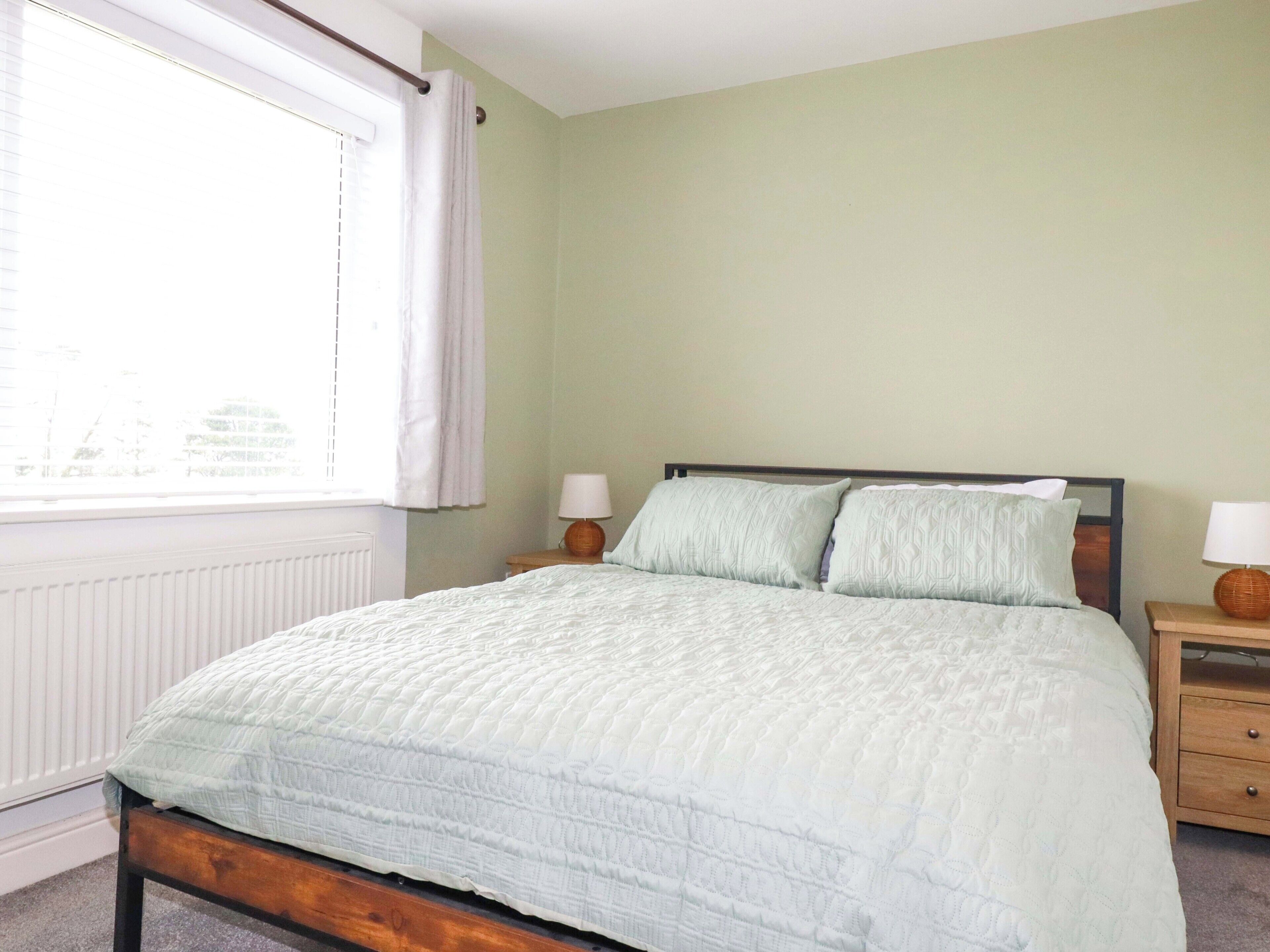 Une chambre à coucher avec un lit, deux tables de chevet, une fenêtre avec des rideaux et un radiateur.