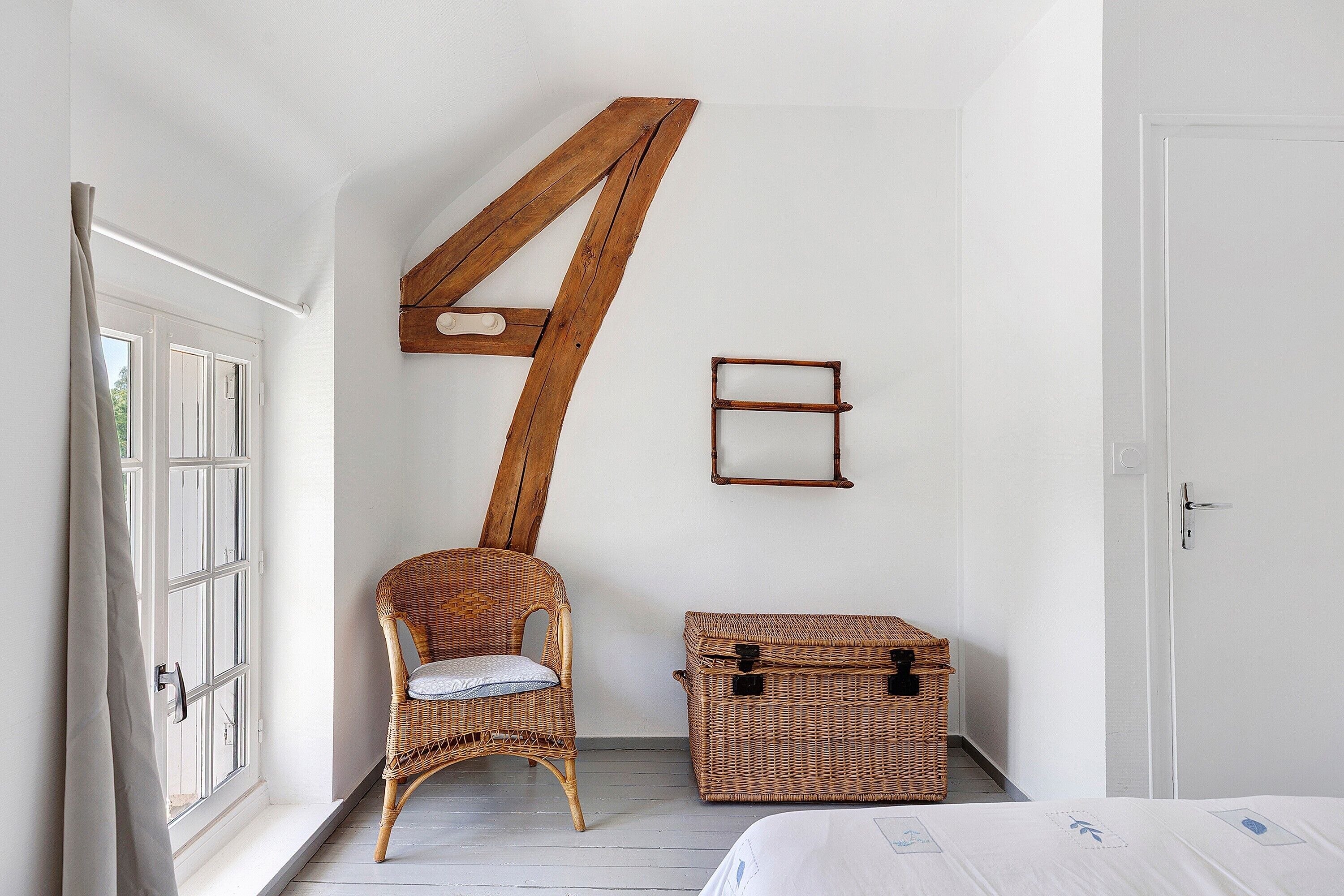 Une pièce comprenant une chaise en bois, une malle en osier et un lit recouvert d’une courtepointe blanche.