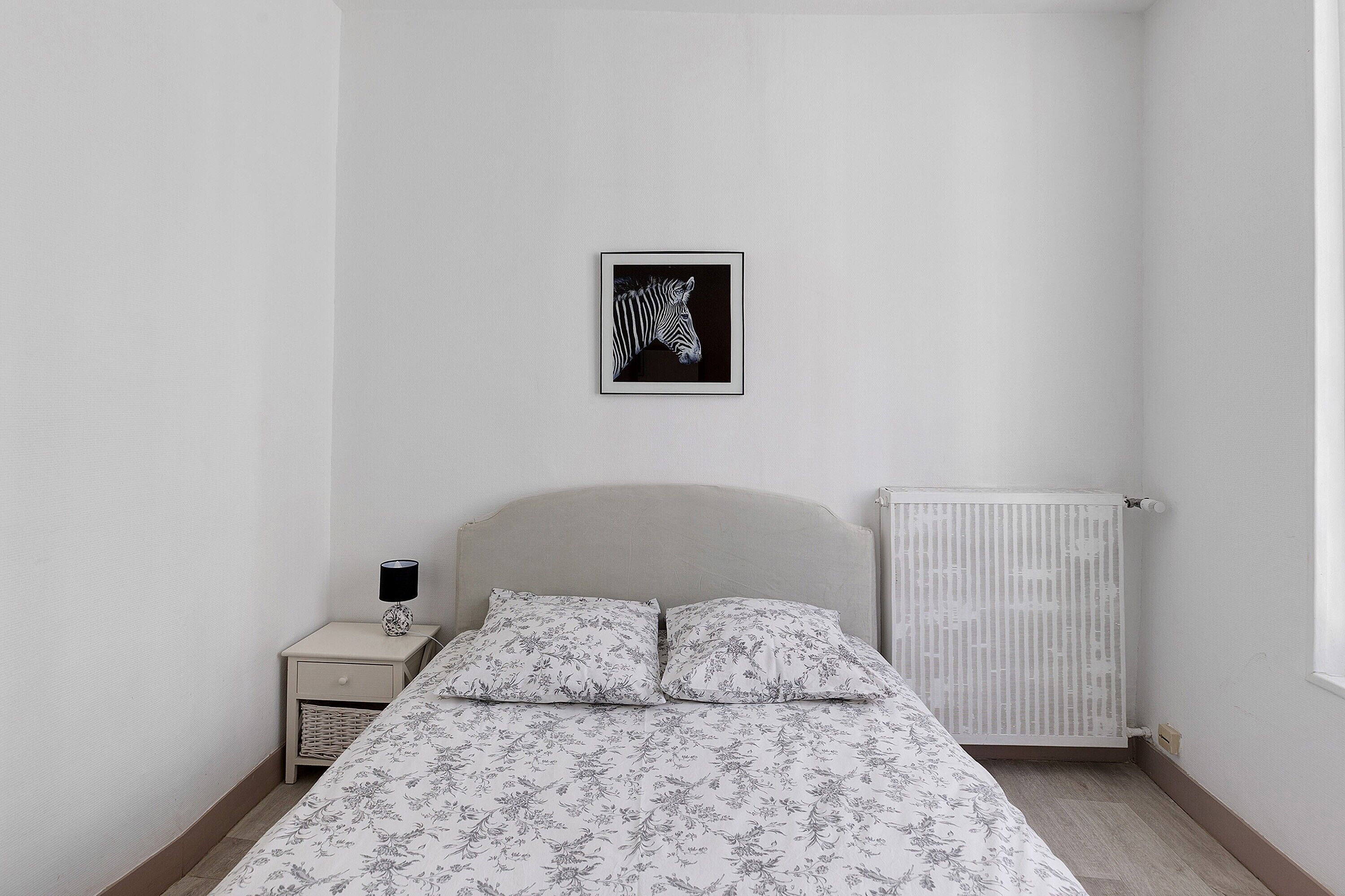 Un dormitorio con cama, mesita de noche, radiador y un cuadro en la pared.