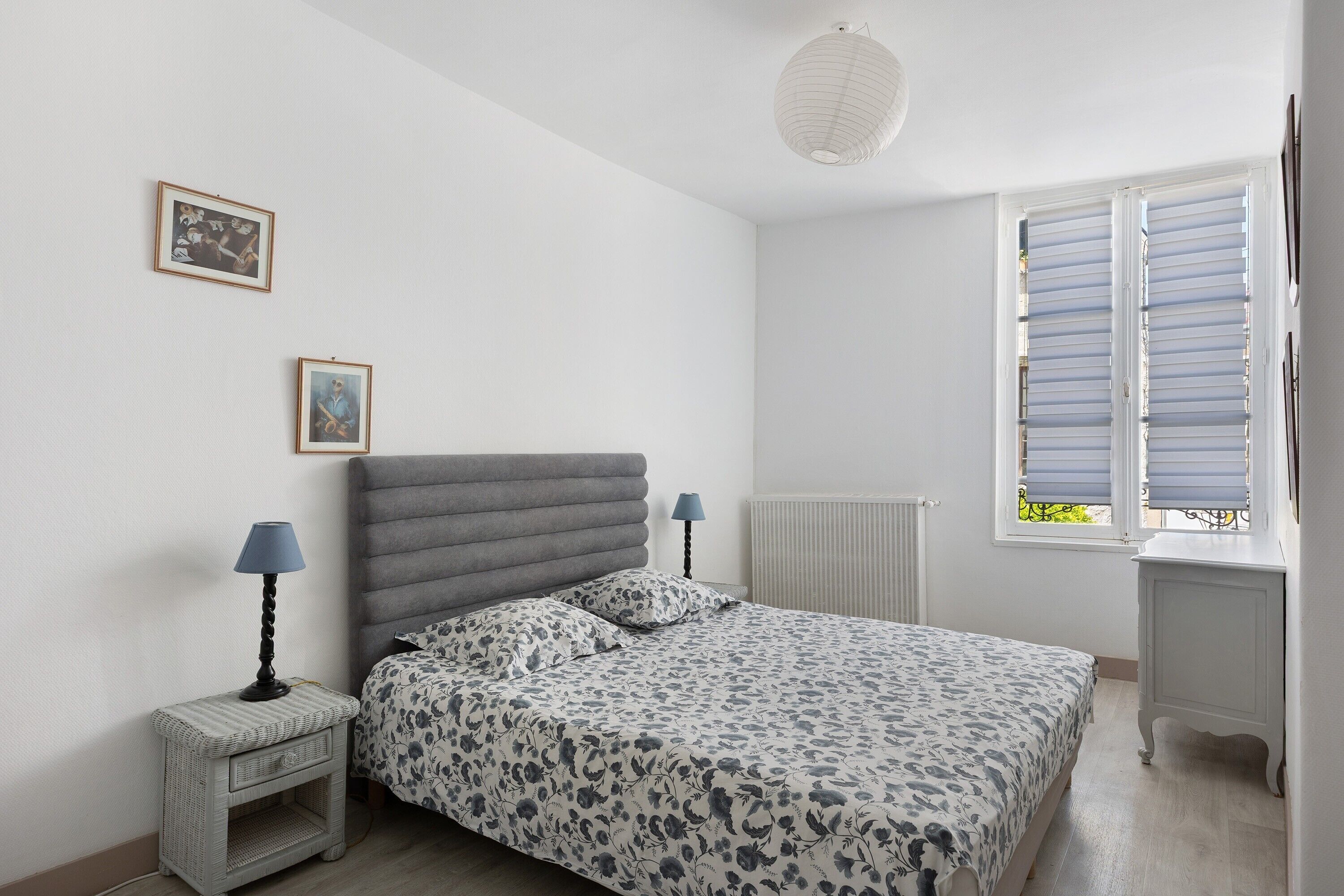 A bedroom with a bed, bedside tables, a radiator, and a window with shutters.