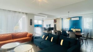 A modern living room with a sofa, ottoman, and coffee table, a dining area with chairs and a table, and a kitchen in the background. - Villa Exclusive 10pers, Garages, 10 min Cathedral (Strasbourg)