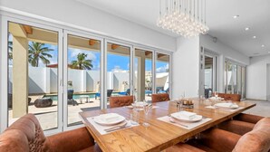 A spacious dining area with a large wooden table, leather chairs, and a view of a pool and palm trees through large glass doors. - LUX 2BR Pool 5mins Beach | Villa Ocean by Bocobay (Noord)