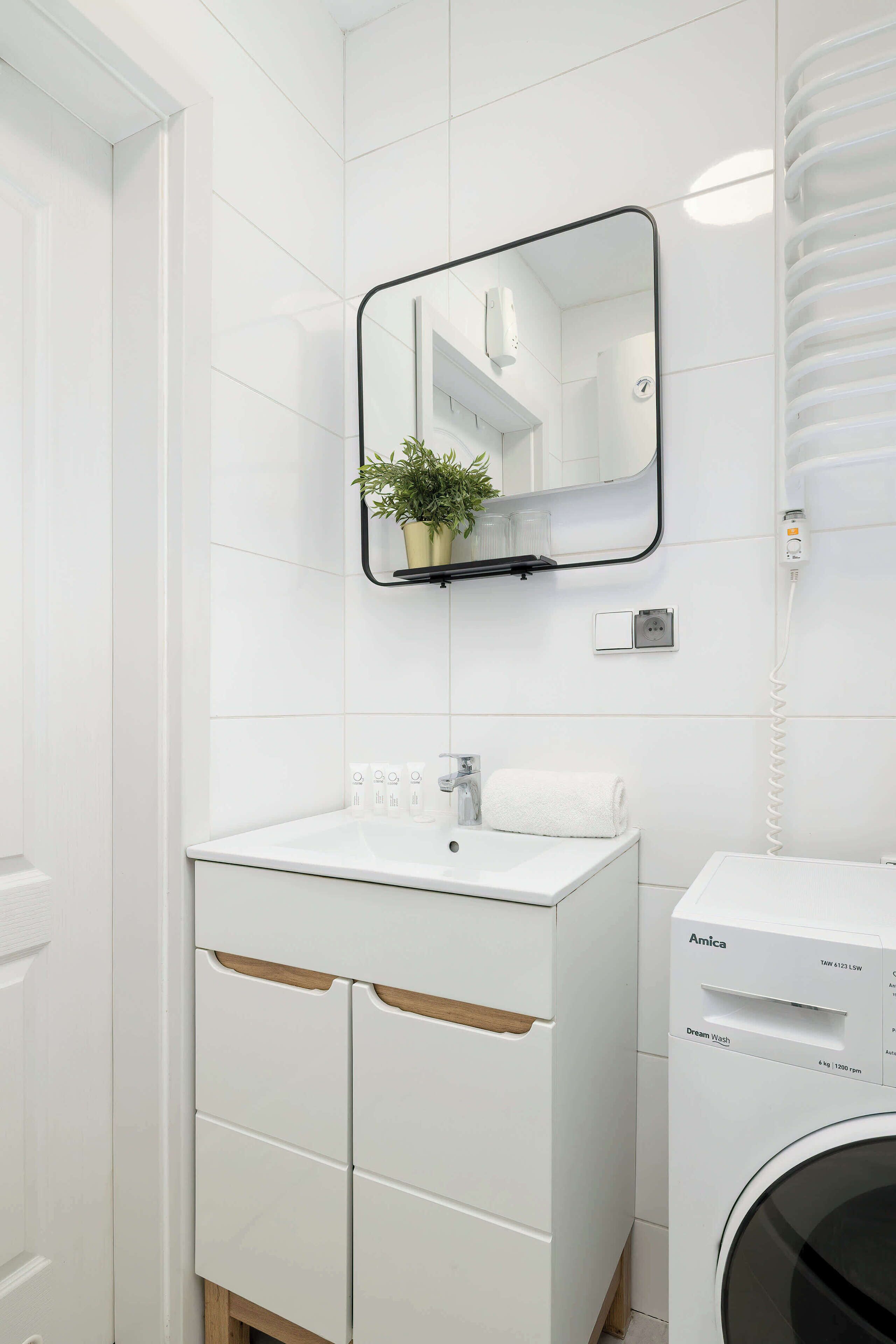 A modern bathroom with white tiles, a sink with a mirror above it, a potted plant, a towel, and a washing machine.