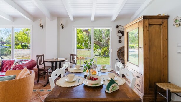 A dining area with a wooden table set for four, surrounded by white chairs, with a view of greenery outside. - Holiday Home 'Tenuta Lu Mimmu' with Private Terrace, Wi-Fi and Air Conditioning (Matino)